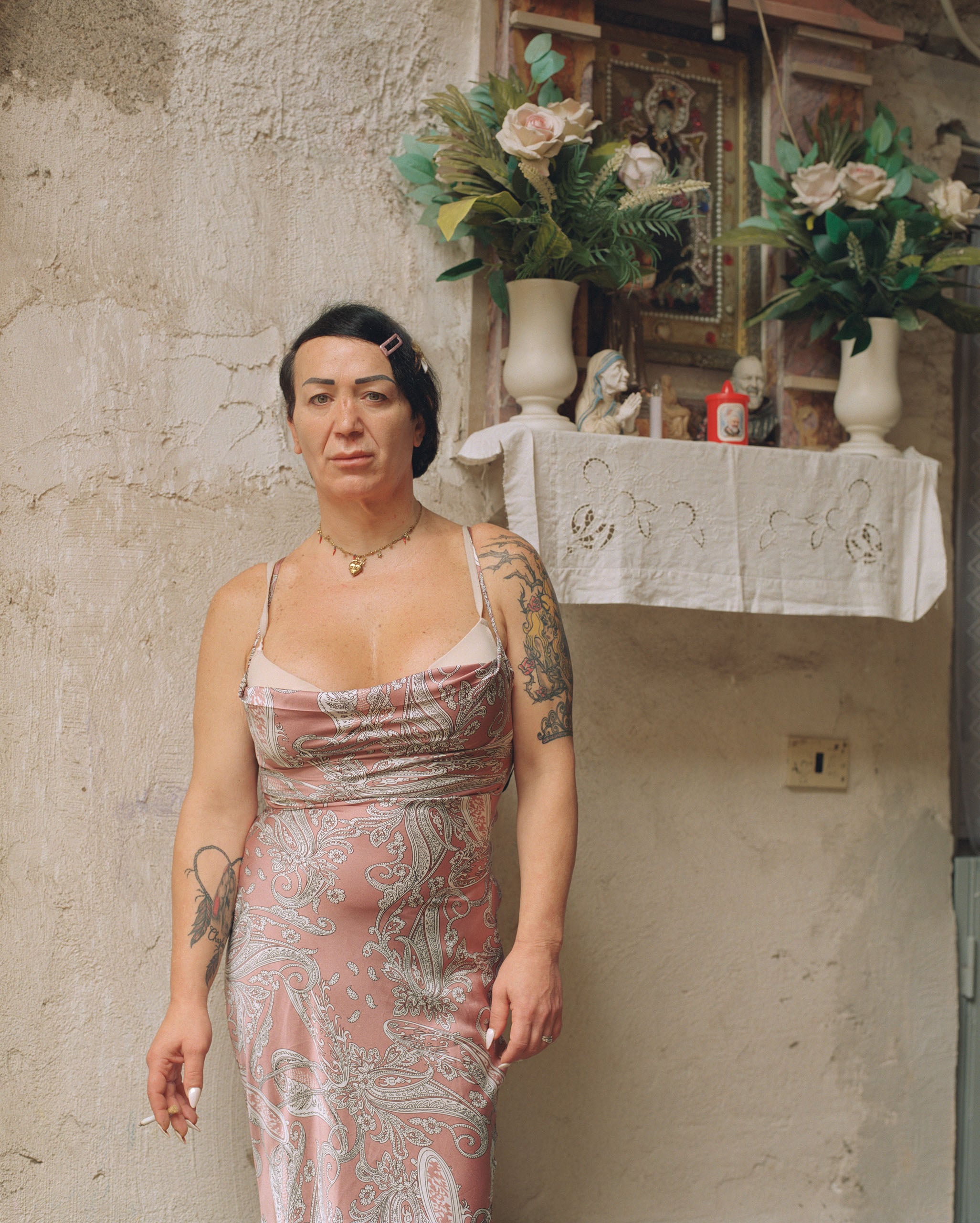 A person with dark hair and tattoos stands in a pink paisley dress, holding a cigarette, in front of a wall-mounted religious shrine with flowers.
