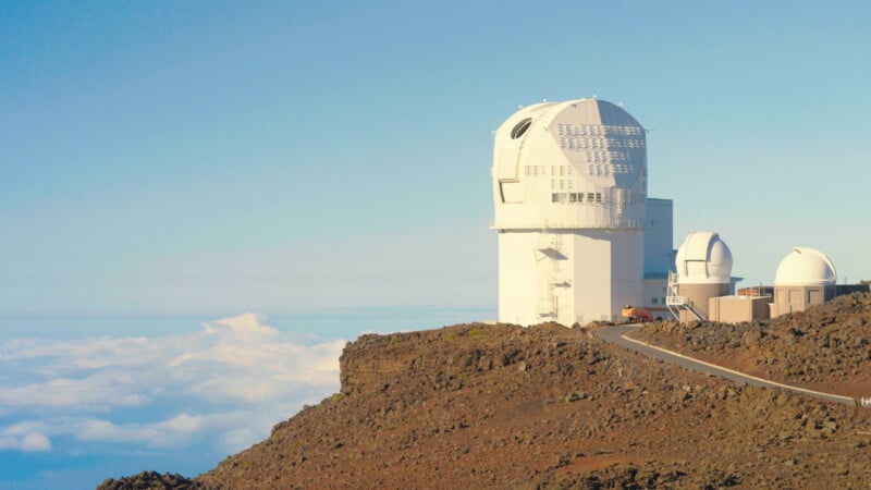 A large white observatory dome stands on a rocky, volcanic ridge above a layer of clouds under a clear blue sky.