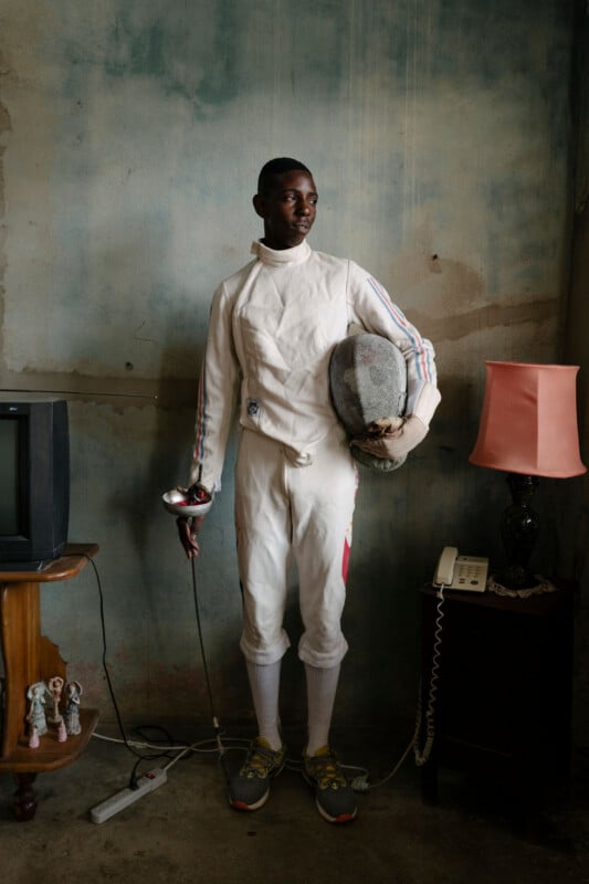 A young fencer in a white uniform holds a mask and foil while standing in a room with a television, a lamp, and an old telephone.