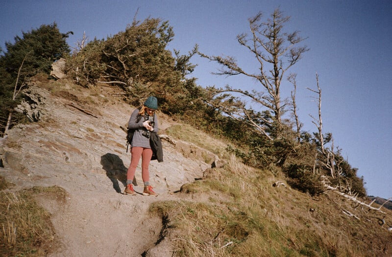 A person wearing a beanie, grey sweater, and reddish leggings stands on a dirt trail on a hillside, looking down at a camera.