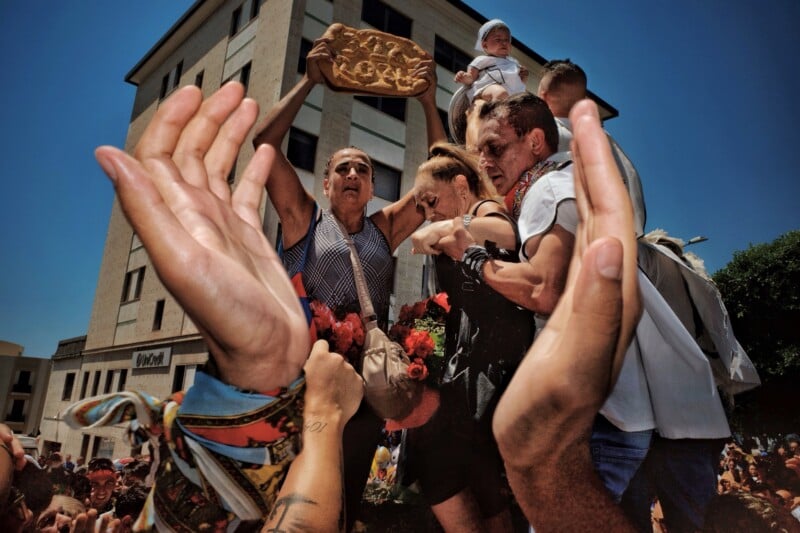 A low-angle shot shows a crowd of people, with hands raised in the foreground, surrounding a woman holding a large, decorated loaf of bread above her head.