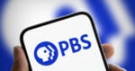 A hand holds a smartphone displaying the blue PBS logo on a white screen against a blurred blue and white background.
