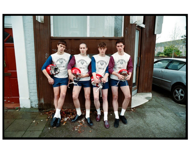 Four young men stand in a row against a wooden wall, wearing matching "Black River Falls Phys. Ed." shirts, blue shorts, and loafers, each holding a red football helmet.