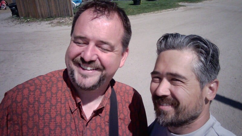Two men with beards pose for a selfie outdoors on a sunny day.