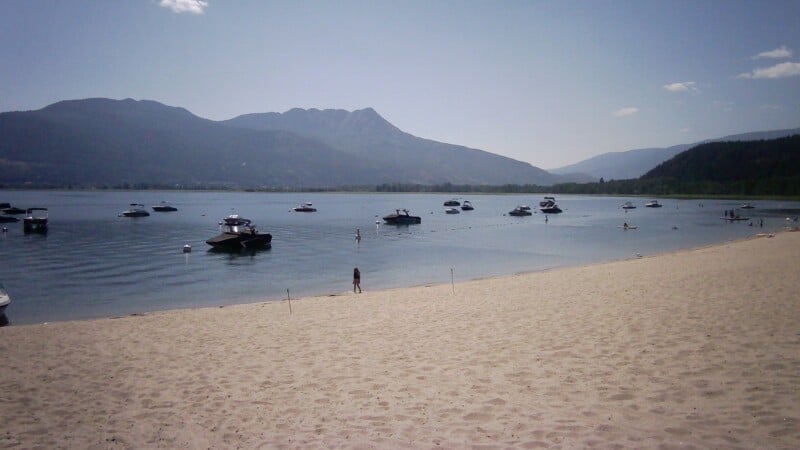 A sandy beach borders a calm lake with numerous motorboats anchored offshore under a clear blue sky and distant mountains.