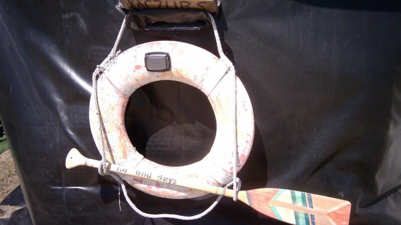 A weathered life ring hangs against a black background, with a wooden paddle featuring a painted geometric design resting horizontally across it.