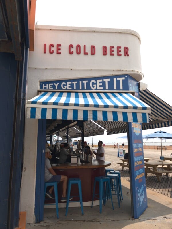 An outdoor bar with a blue and white striped awning features a sign reading "ICE COLD BEER" and "HEY GET IT GET IT" above a counter where people are sitting on blue stools.