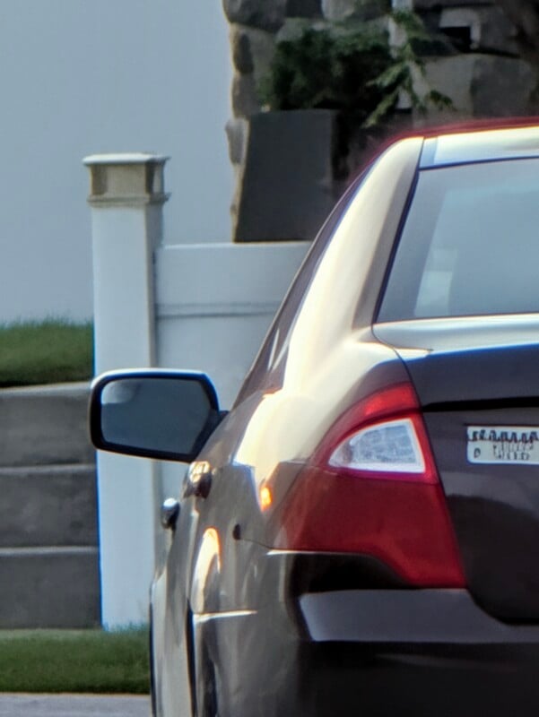 The rear corner of a dark-colored sedan is parked next to a white fence post and stone wall.