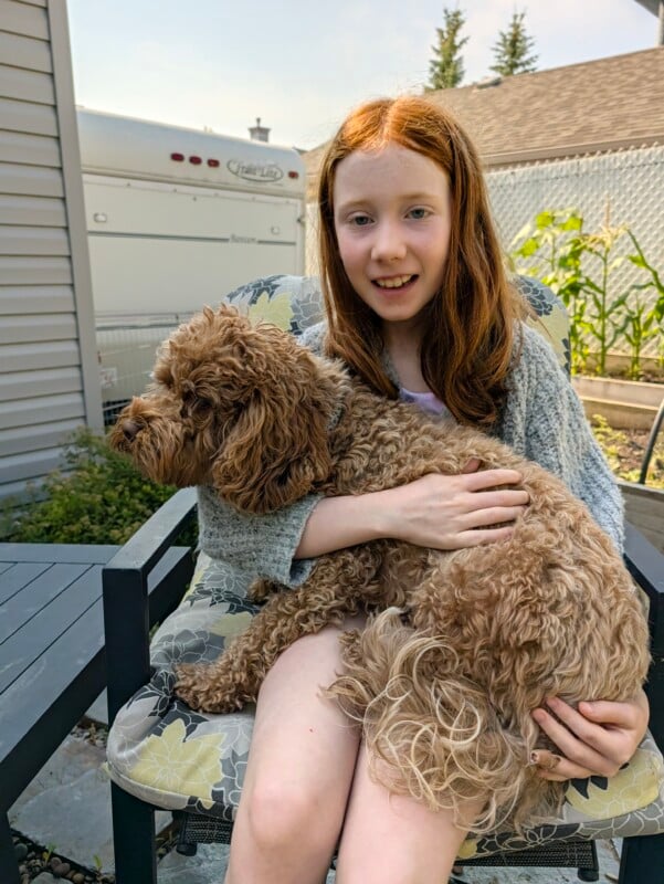 A young girl with long red hair sits in a patterned outdoor chair, smiling while holding a curly, light-brown dog in her lap.