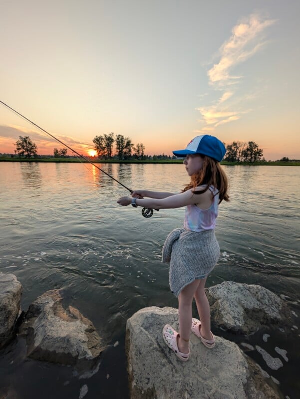 A young girl in a baseball cap and Crocs stands on rocks by a river at sunset, holding a fly fishing rod.