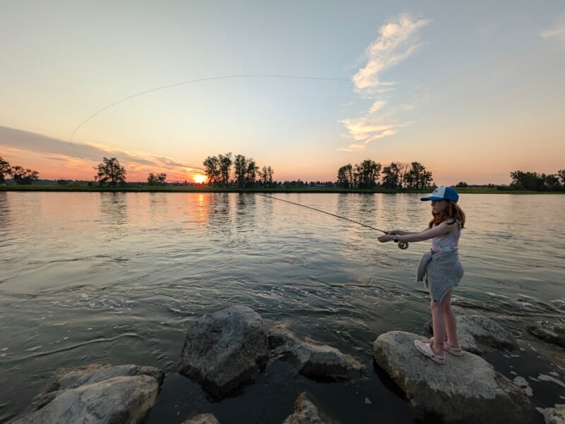 A young girl in a baseball cap and Crocs fly fishes while standing on rocks in a river during a sunset.