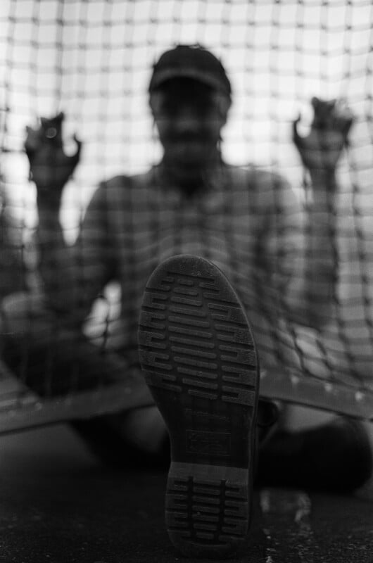 A close-up, low-angle view of a shoe sole with a tread pattern, with a blurred person sitting behind a chain-link fence in the background.