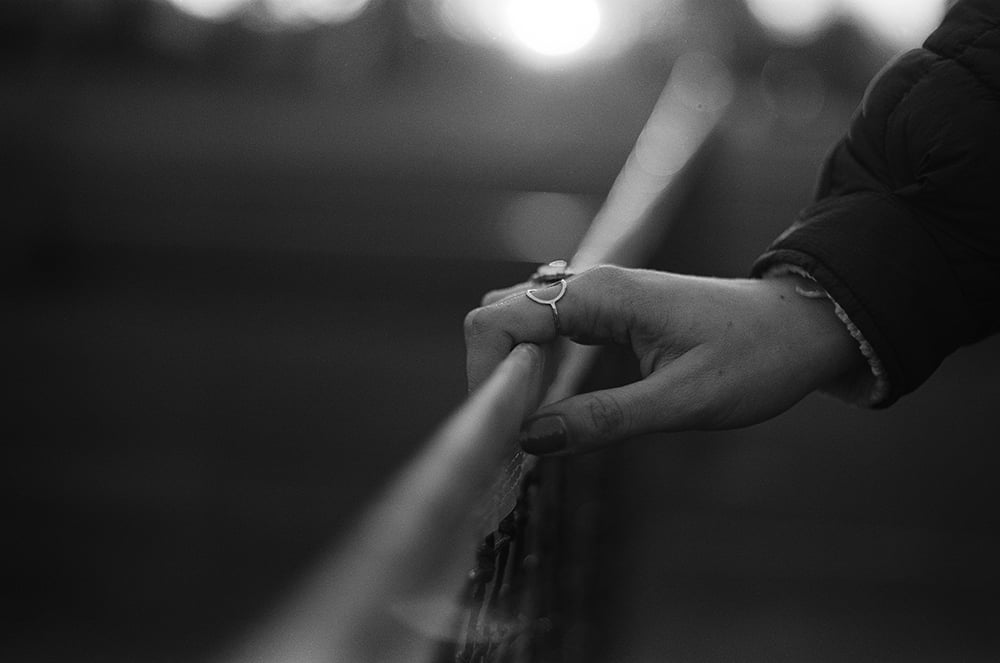 A close-up, black-and-white shot of a hand resting on a metal railing, wearing a thin ring with a small circular detail.