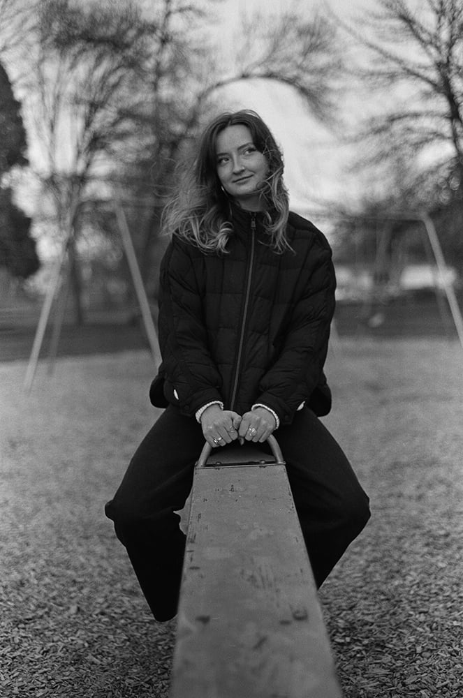 A young woman with wavy hair sits on a seesaw in a park, wearing a black quilted jacket and dark pants.