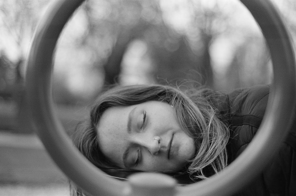 A person with closed eyes rests their head against a circular metal frame in an outdoor setting.