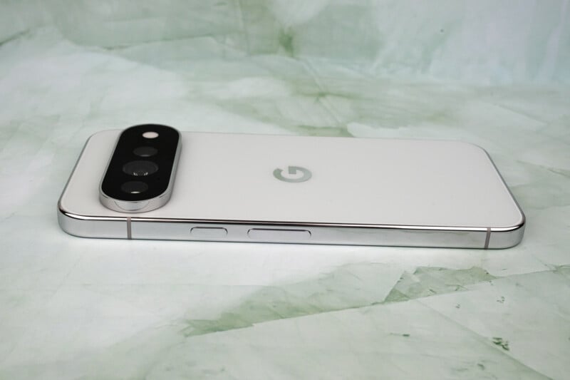 A white smartphone with a prominent, pill-shaped camera module on the back lies on a light green, marbled surface.