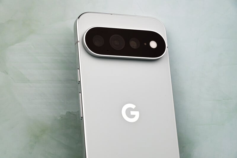 A close-up, angled view of the back of a light gray smartphone featuring a prominent, horizontal, pill-shaped camera module with three lenses and a flash, and a white "G" logo centered on the body.