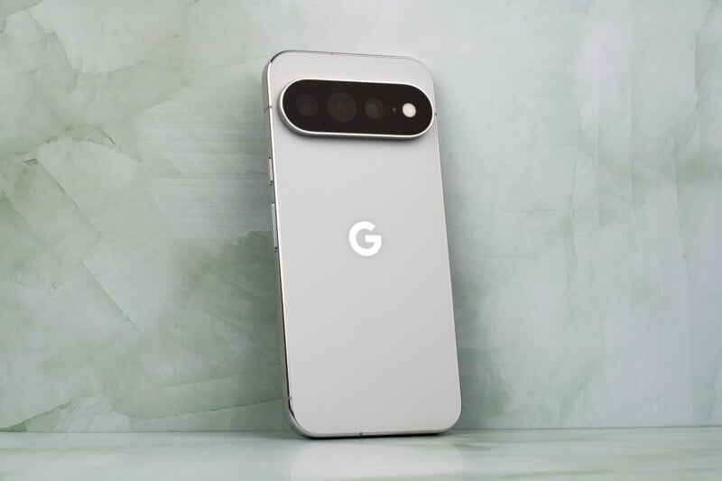 A silver smartphone with a prominent horizontal camera bar on its back stands upright against a pale green marble background.