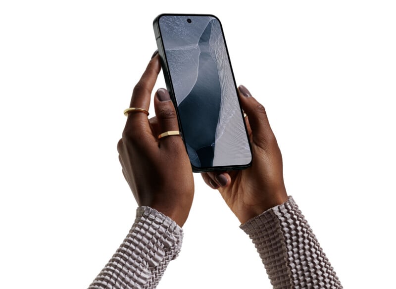 A pair of dark-skinned hands, wearing gold rings and a textured long-sleeved top, hold a smartphone displaying an abstract, monochromatic landscape wallpaper.
