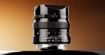 A black camera lens stands upright against a gradient brown background, showing aperture and focus distance markings on its barrel.