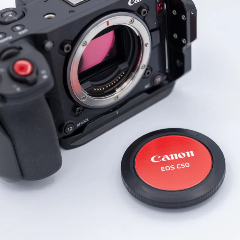 A black Canon cinema camera body with its lens mount exposed sits next to a circular red and black body cap labeled "Canon EOS C50" on a white surface.