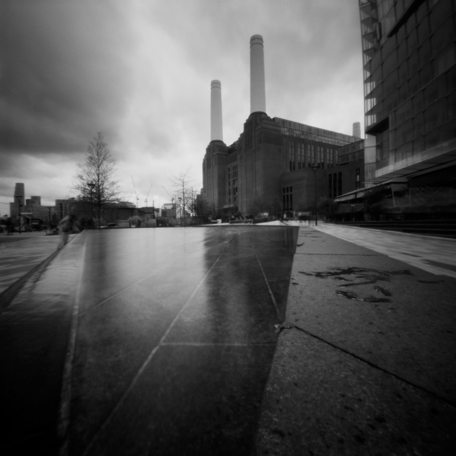 A black-and-white low-angle shot of a large industrial building with two tall chimneys behind a reflective stone plaza.