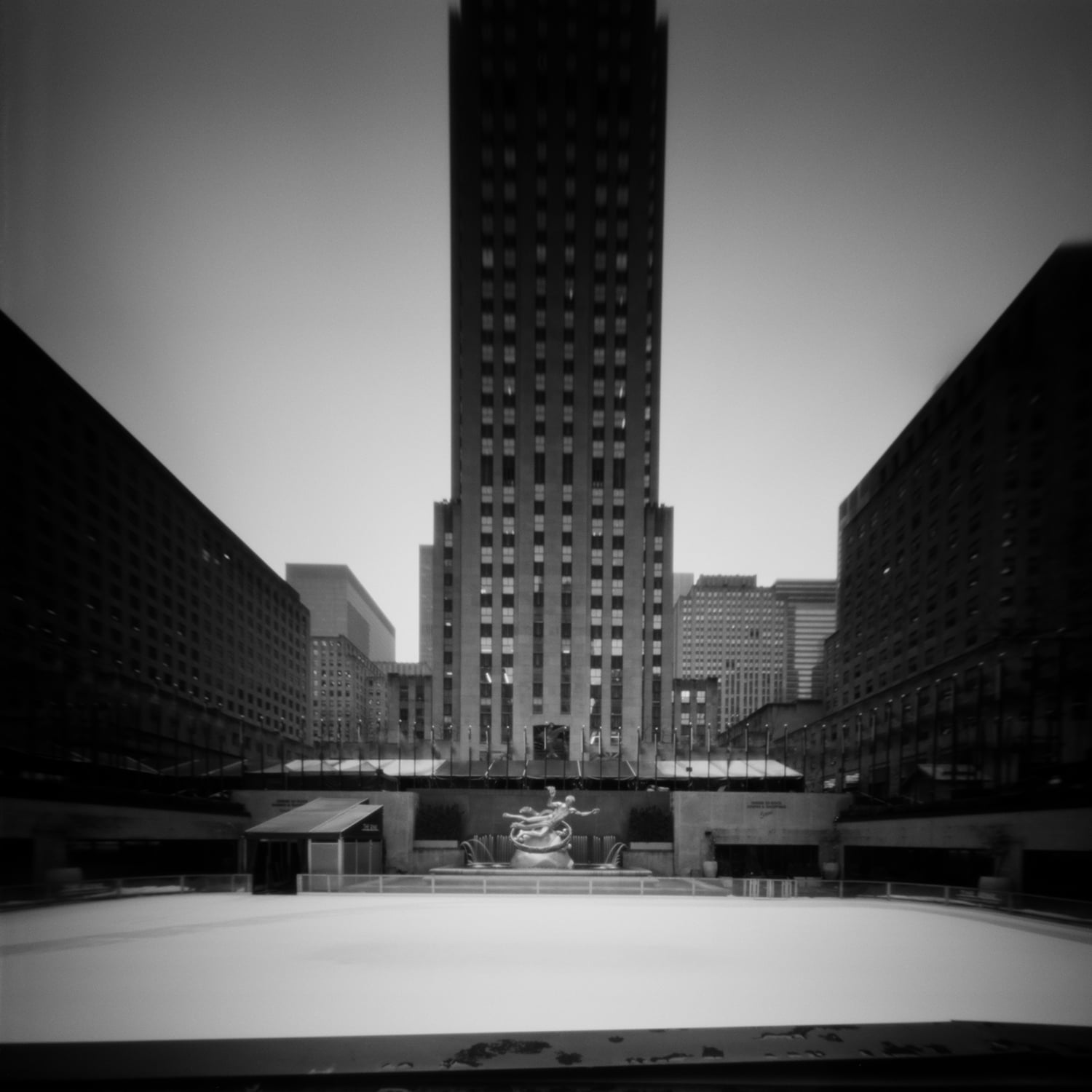 A black-and-white, low-angle view of a tall skyscraper rising above a large, empty ice rink with a central fountain statue.