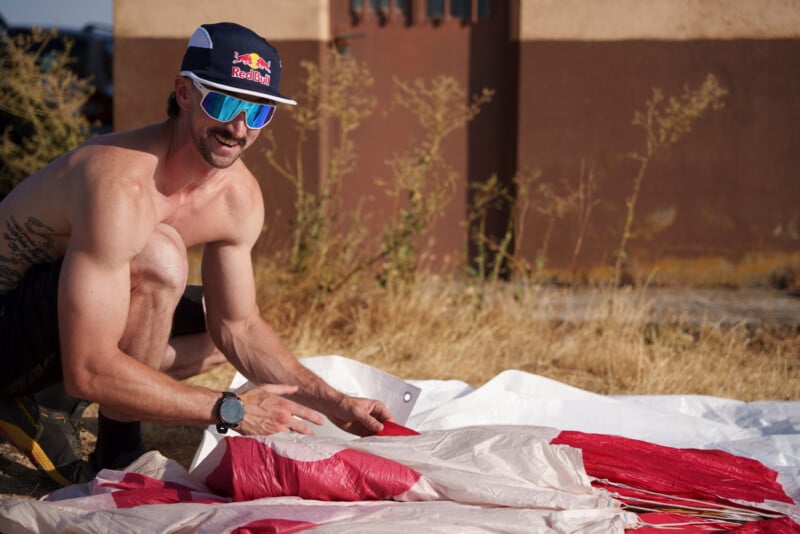 A shirtless man wearing a Red Bull cap and blue-tinted sunglasses kneels on the ground, folding a red and white parachute.