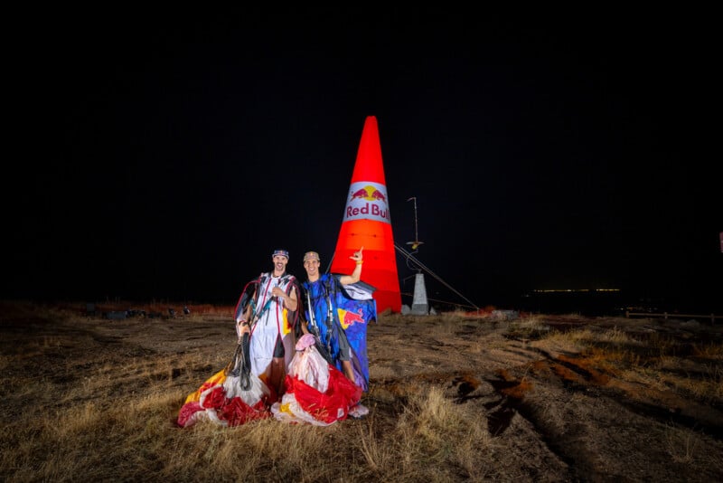 Two skydivers in wingsuits stand in a field at night in front of a large, illuminated orange Red Bull pylon.
