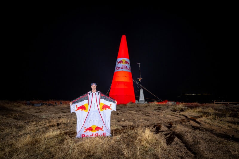 A person wearing a Red Bull branded wingsuit stands in a field at night in front of a large, illuminated red inflatable cone.