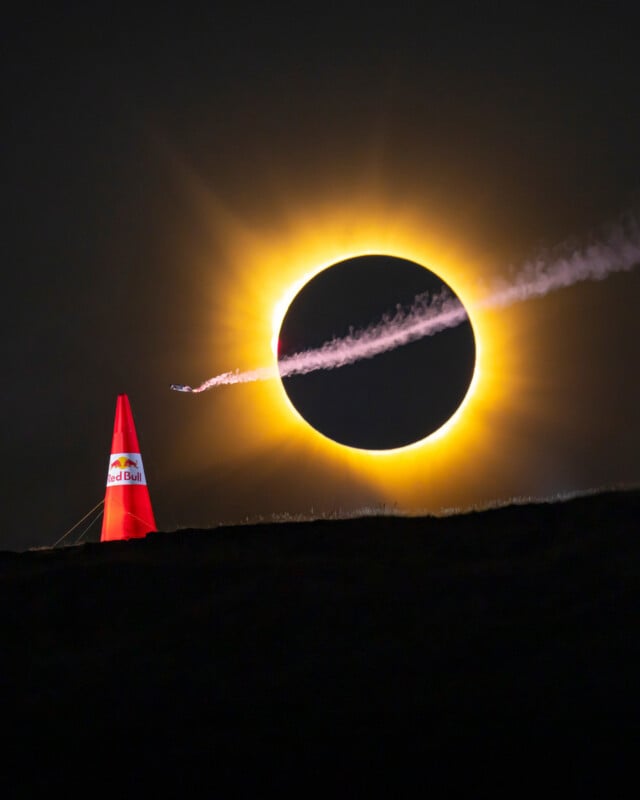 A small airplane with a smoke trail flies past a red Red Bull pylon in front of a total solar eclipse.