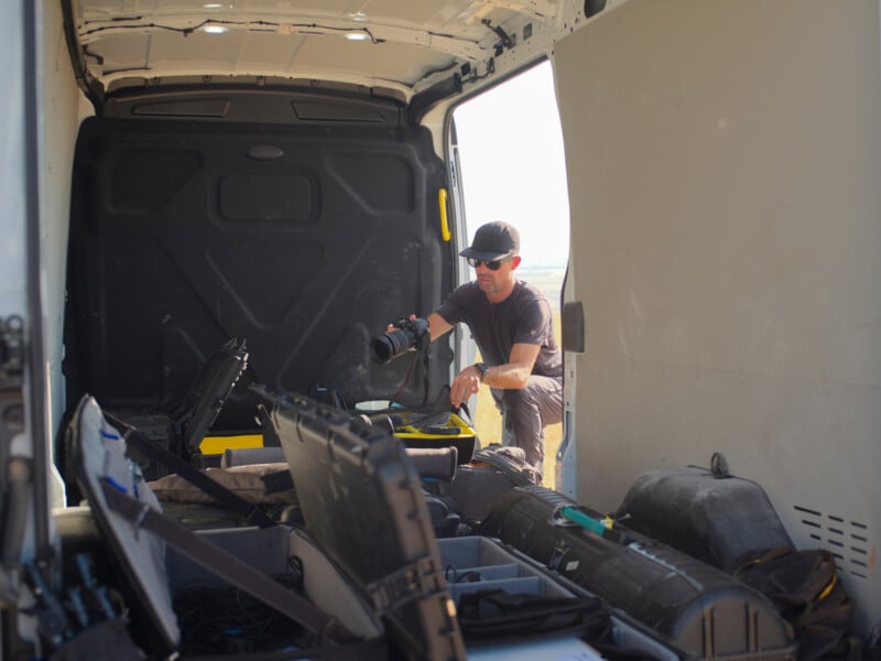 A man wearing a cap and sunglasses kneels inside the back of a van, holding a camera while surrounded by open equipment cases.