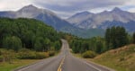 A paved road stretches into the distance toward a range of large, rugged mountains under a cloudy sky, flanked by dense green forests.