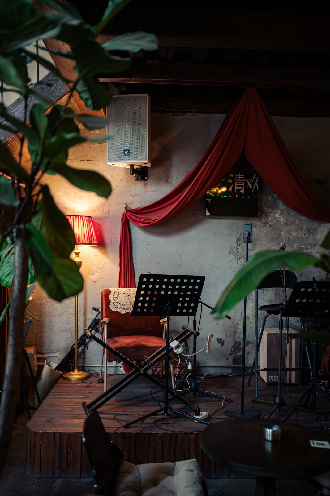A small wooden stage features a music stand, a red velvet chair with a lace doily, a floor lamp, and a speaker mounted on a textured wall draped with red fabric.
