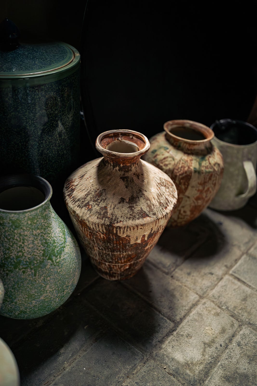 A collection of various ceramic vases and pots with different textures and glazes sits on a stone-tiled floor.