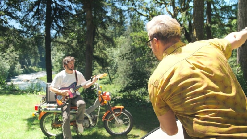 A man in a white t-shirt plays an electric guitar while sitting on an orange moped, as another man in a yellow patterned shirt watches.