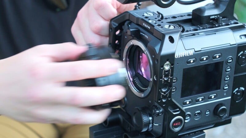 A person's hands are attaching a lens to the front of a Fujifilm cinema camera equipped with a Tilta cage.