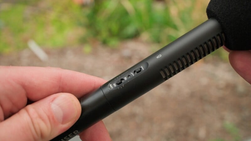 A hand holds a black shotgun microphone, showing a switch set to the "ON" position and a low-cut filter icon.