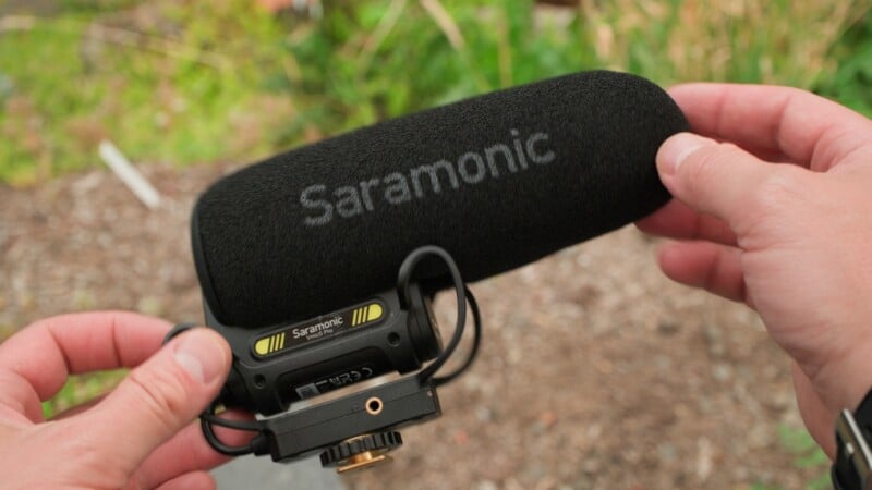 A person holds a black Saramonic Vmic5 Pro shotgun microphone with a foam windscreen against a blurred outdoor background.