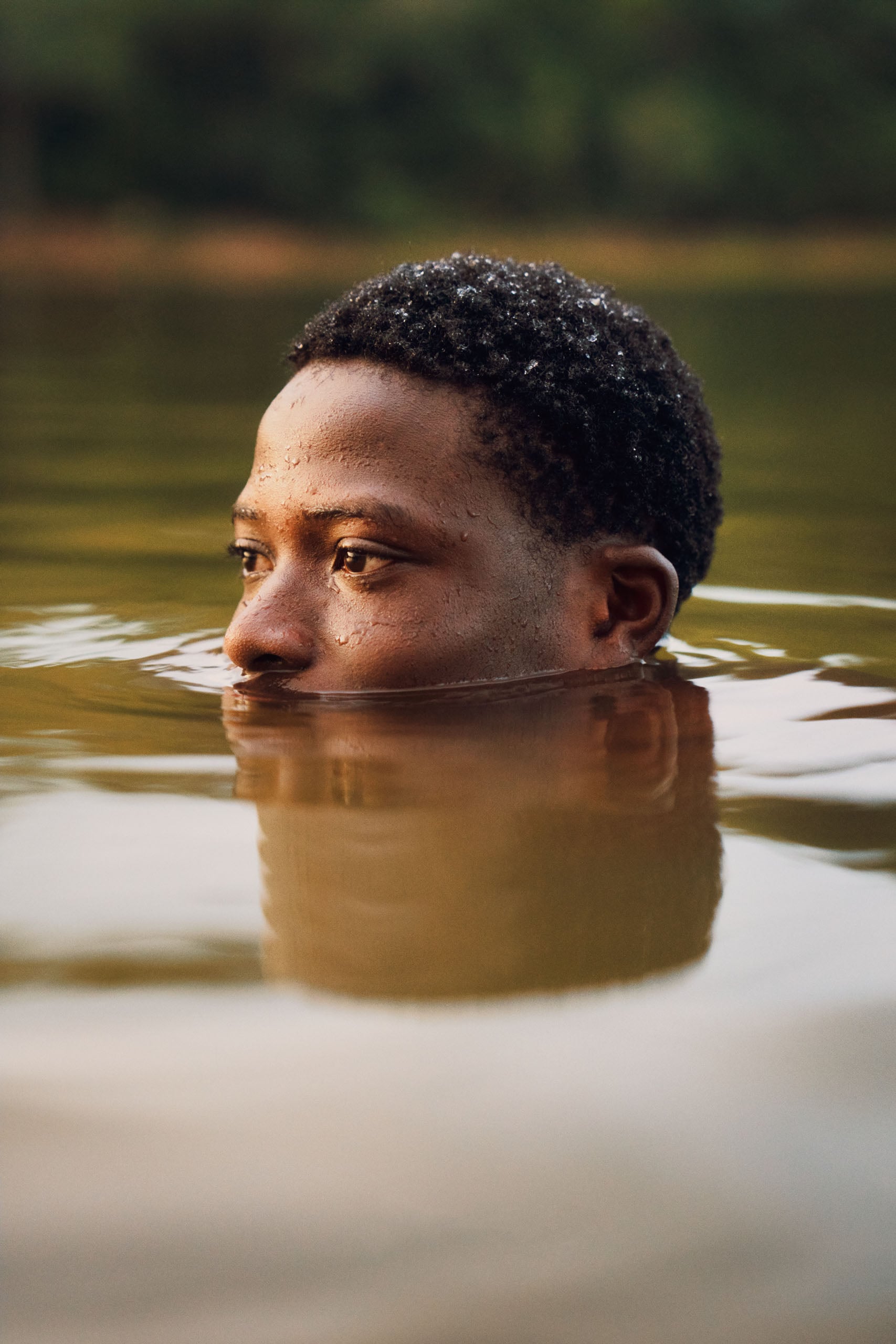 A person with short, dark, textured hair is partially submerged in calm, murky water, looking off to the side.