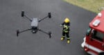 A drone hovers in the air while a firefighter in uniform operates it with a remote control near a red fire truck.