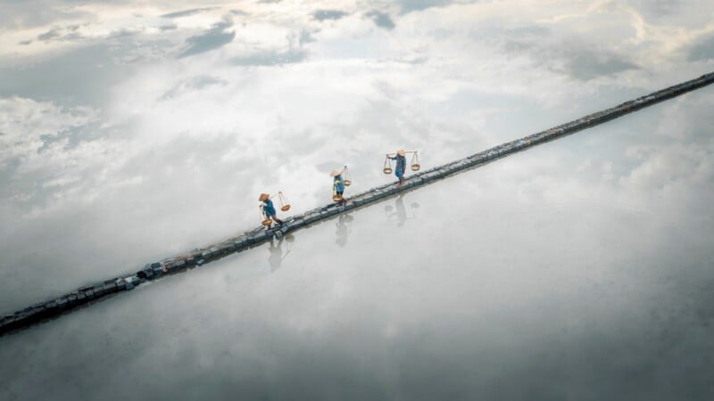 Three people wearing conical hats and carrying baskets on shoulder poles walk in a line across a narrow stone path over water.