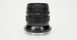 A black manual focus camera lens stands upright against a plain white background, showing aperture and distance markings.