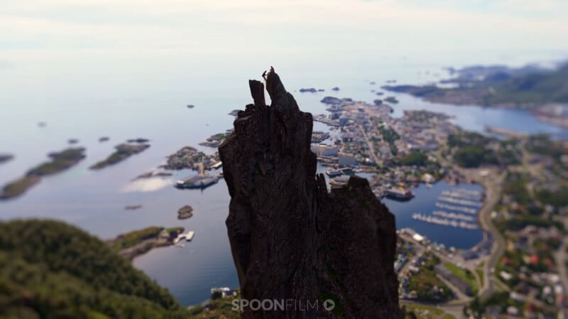 Two climbers stand on the narrow, jagged peak of a mountain overlooking a coastal town and harbor.