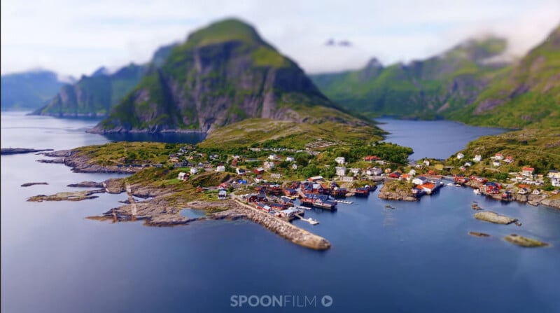 A small coastal village with red and white houses sits on a rocky peninsula surrounded by water and dramatic green mountains.