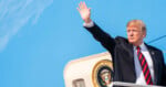 Donald Trump waves his hand while standing in front of the Presidential Seal on the side of an aircraft against a bright blue sky.