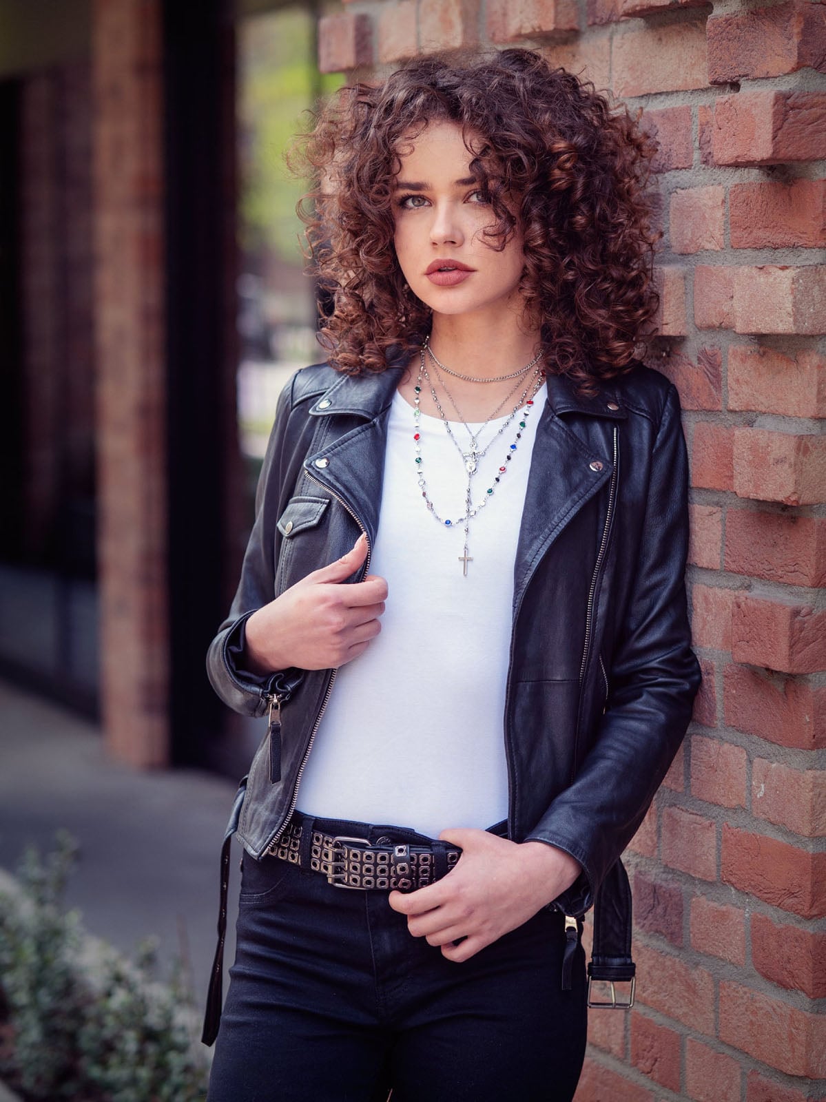 A young woman with curly brown hair wearing a black leather jacket, white t-shirt, and a studded belt leans against a brick wall.