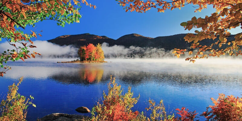A small island with vibrant autumn foliage sits in the middle of a calm, blue lake surrounded by mist and forested mountains.