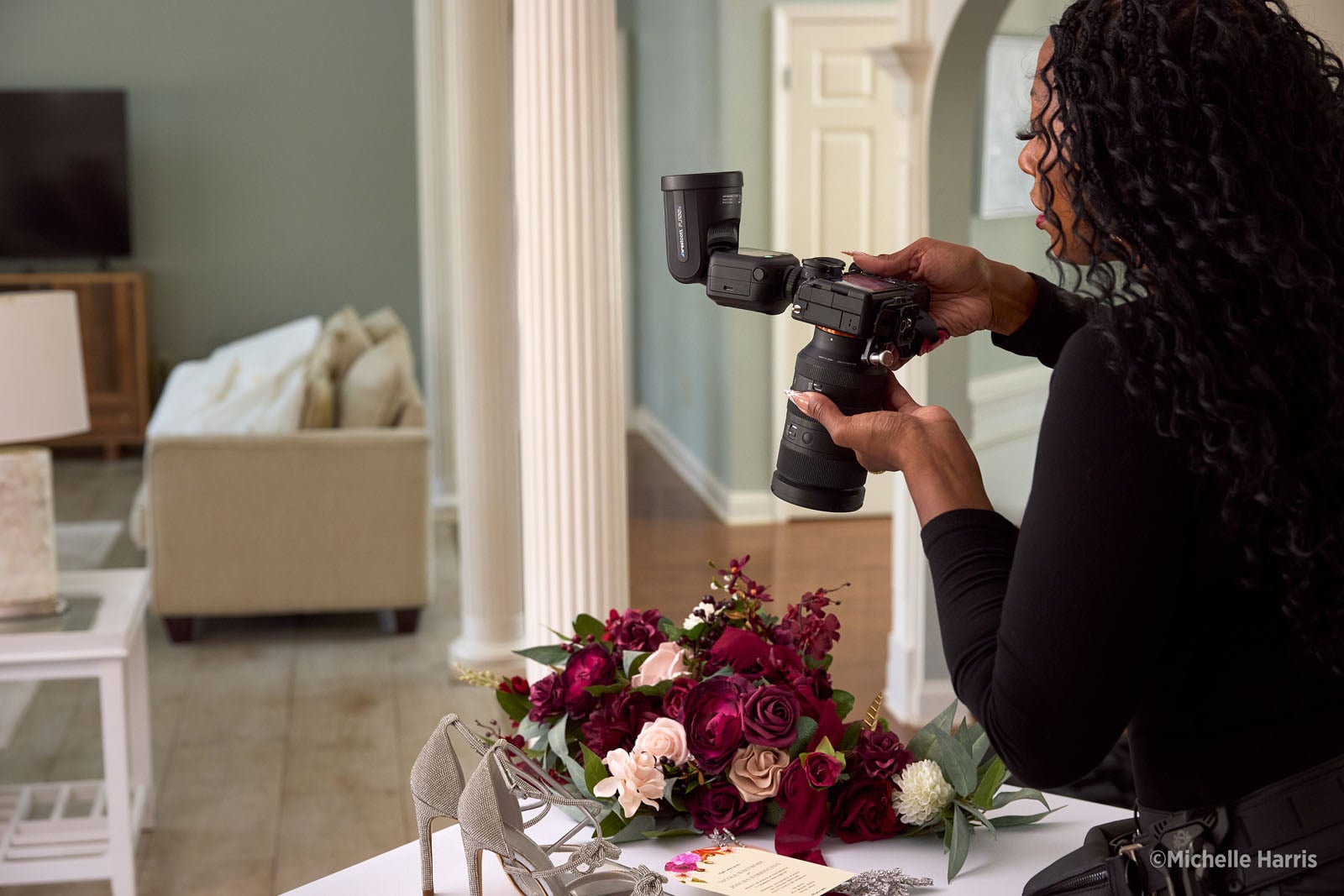 A photographer with long dark hair holds a camera with an attached flash, aiming it at a floral arrangement and a pair of silver heels on a table.
