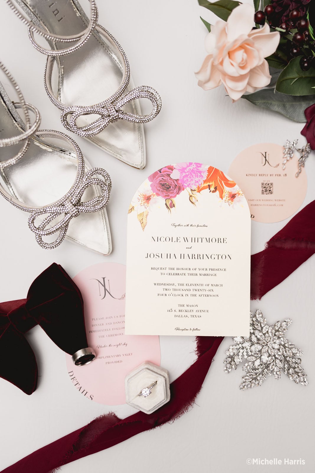 A flat-lay of wedding stationery featuring an arched invitation for Nicole Whitmore and Josuha Harrington, surrounded by silver heels, a velvet bow tie, a ring in a box, floral accents, and burgundy ribbon.